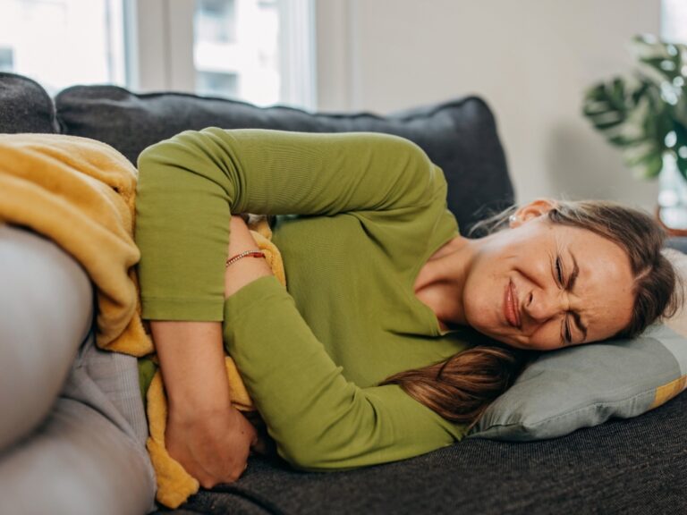 Frau liegt mit schmerzverzerrtem Gesicht auf dem Sofa und hält sich den Bauch – Symbolbild für akute Beschwerden nach Nahrungsmittelunverträglichkeit.