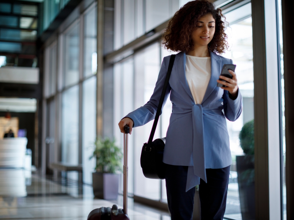 Eine Geschäftsfrau mit Rollkoffer und Schultertasche geht durch die Lobby eines modernen Gebäudes und schaut auf ihr Smartphone. Sie wirkt gut vorbereitet – auch in Bezug auf Ernährung bei Nahrungsmittelunverträglichkeiten während der Geschäftsreise.