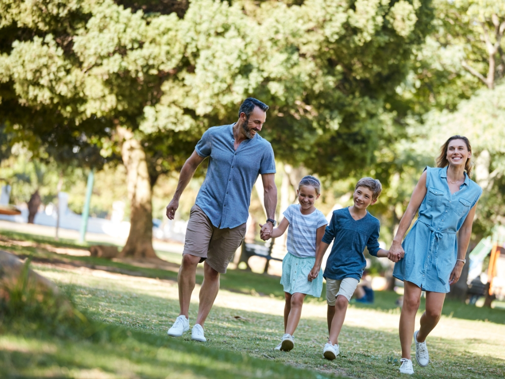Eine fröhliche Familie spaziert an einem sonnigen Tag durch einen Park. Sie wirken aktiv und ausgeglichen – ein Symbol für einen bewussten, genussvollen Lebensstil, bei dem auch Menschen mit Nahrungsmittelunverträglichkeiten ihre Wohlfühlroutinen beibehalten.